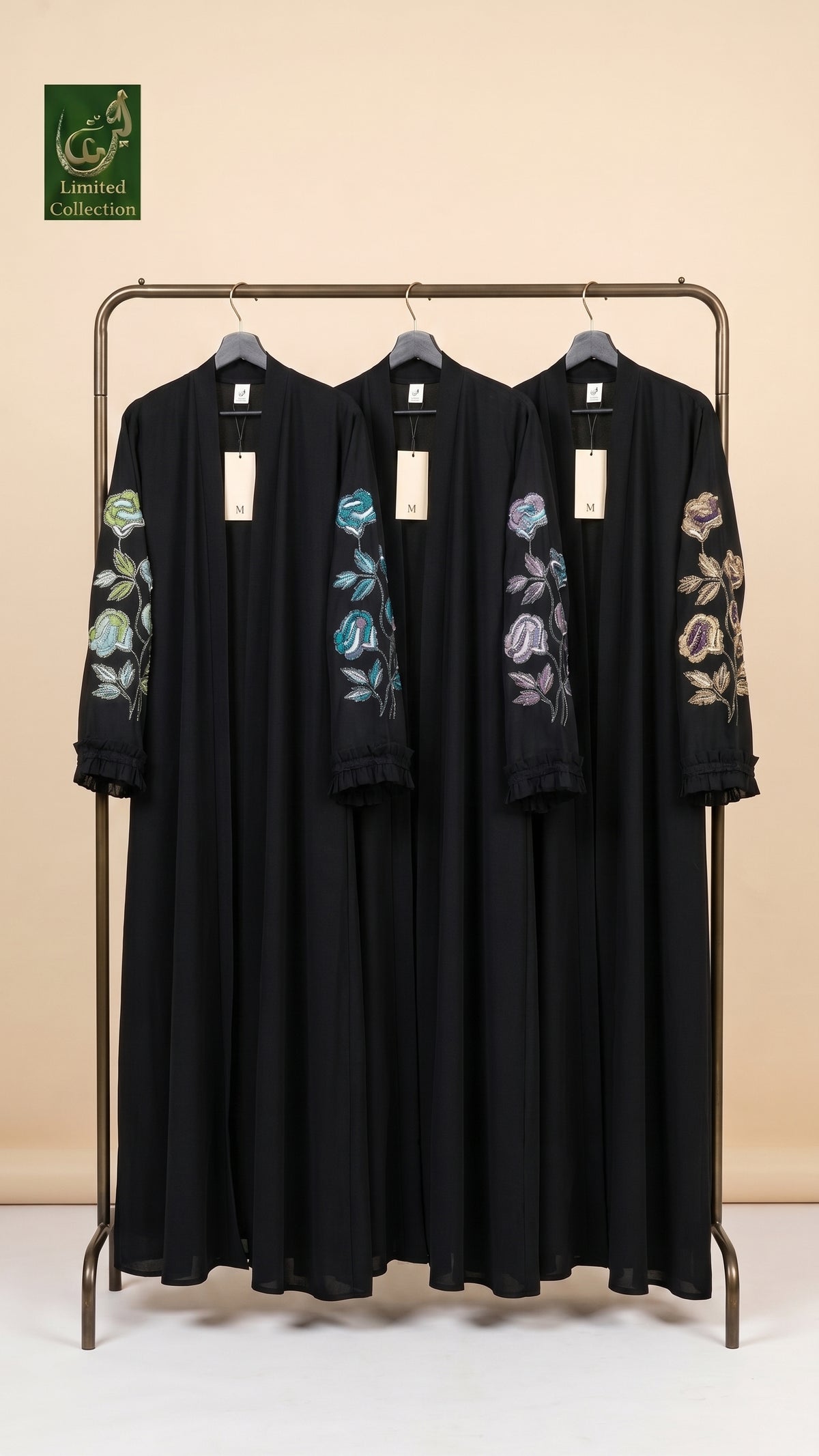 Three black long-sleeve abaya dresses with colorful floral embroidery on the sleeves, hanging on a metal clothing rack against a beige background. A green 'Limited Edition' logo is visible in the top left corner.