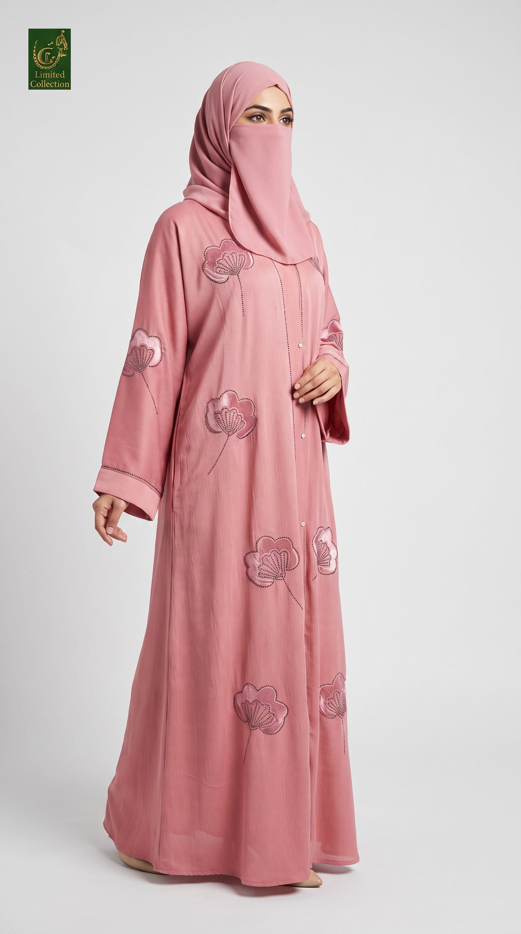 A woman modeling a dusty pink abaya with matching hijab, featuring embroidered floral patterns, set against a plain light gray background.