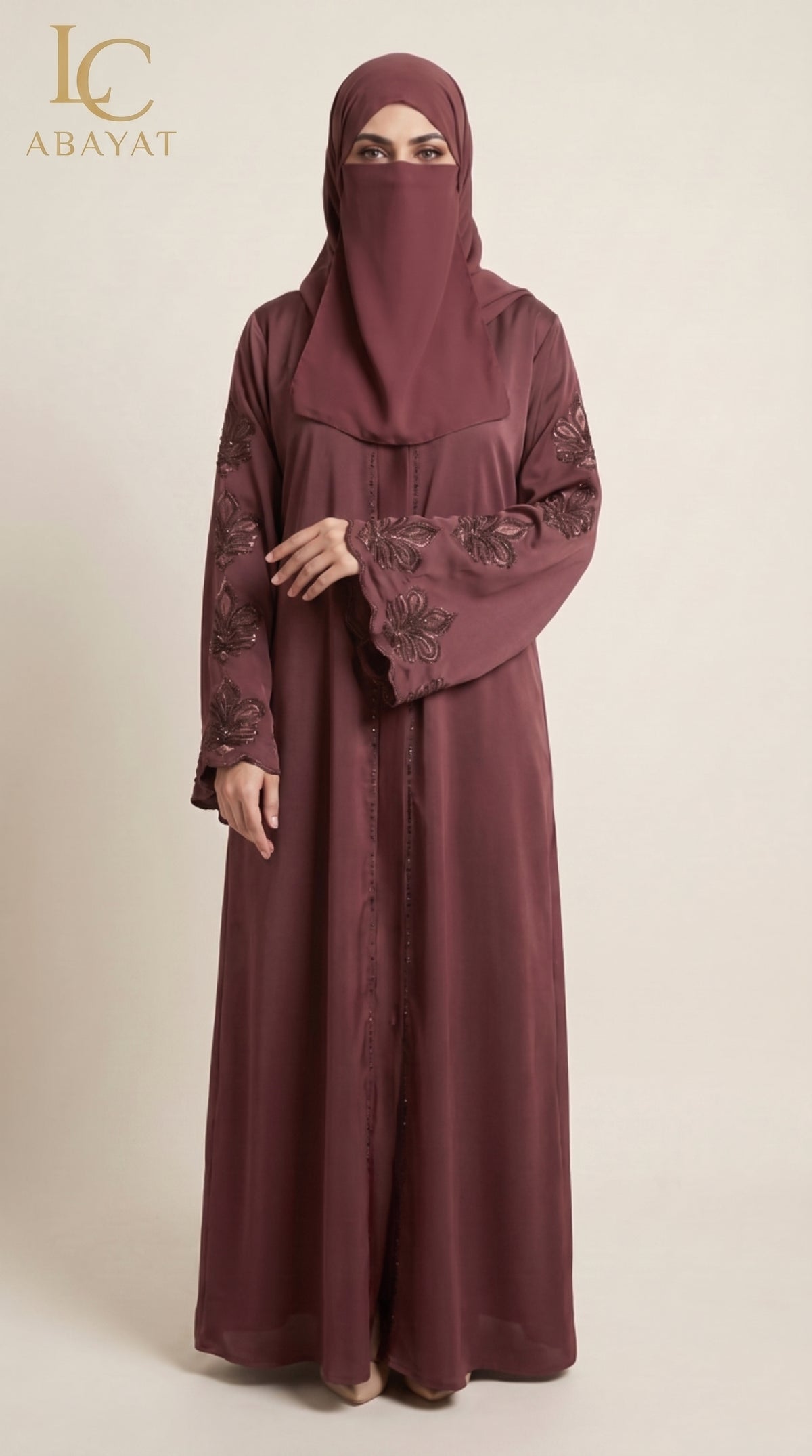 A woman wearing a long maroon abaya with floral embroidery on the sleeves and a matching hijab, standing against a plain beige studio background. The LC Abayat brand logo is visible in the top left corner.