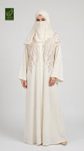 A woman wearing a long cream abaya with intricate lace embroidery on the chest, paired with a matching hijab, standing against a light gray studio background. A green 'Limited Collection Abayat' logo is visible in the top-left corner.
