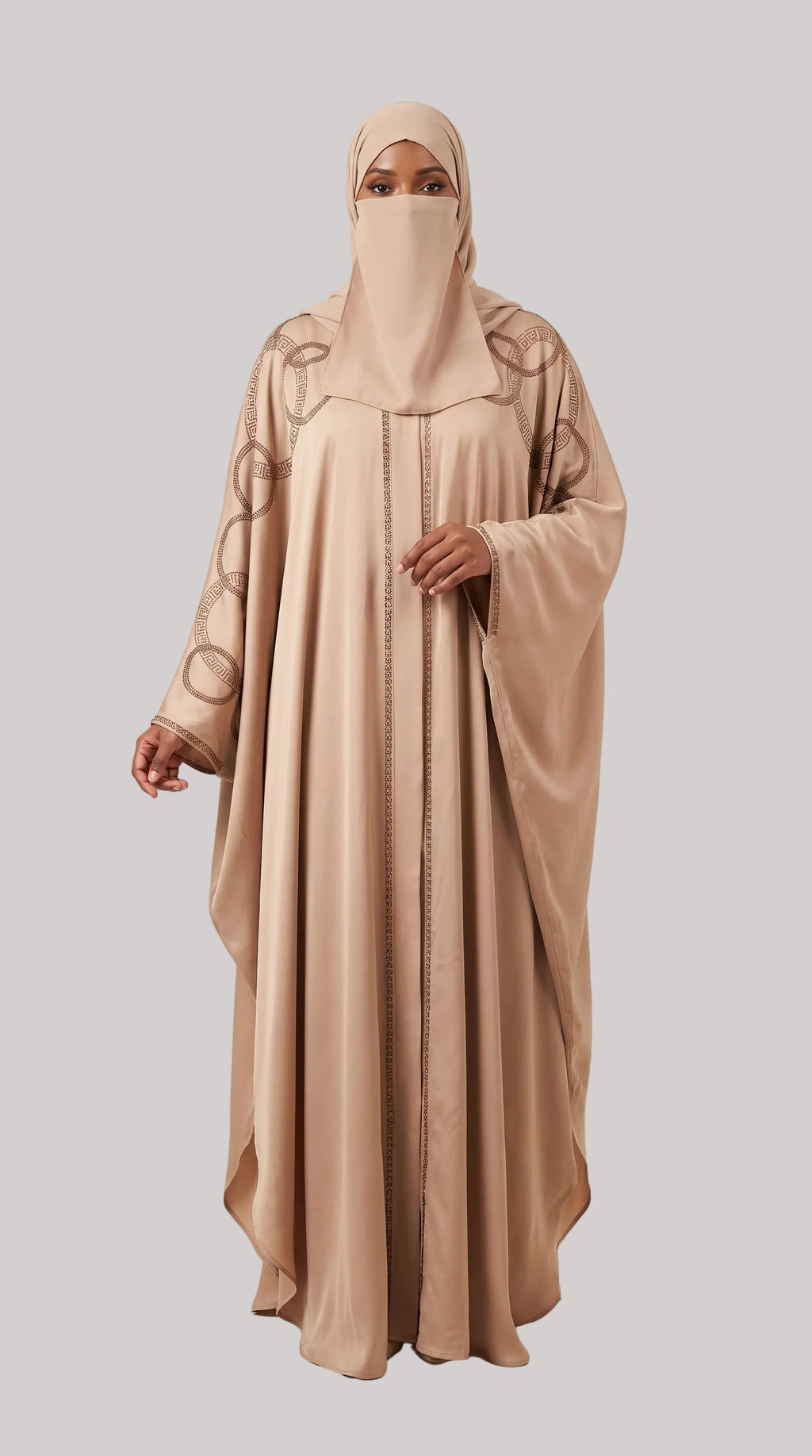 A woman modeling a modest abaya and matching hijab set in a warm beige/tan shade, standing against a plain light beige studio background. The abaya has long flowing sleeves and subtle circular embroidered motifs near the upper sleeves, with a front closure.