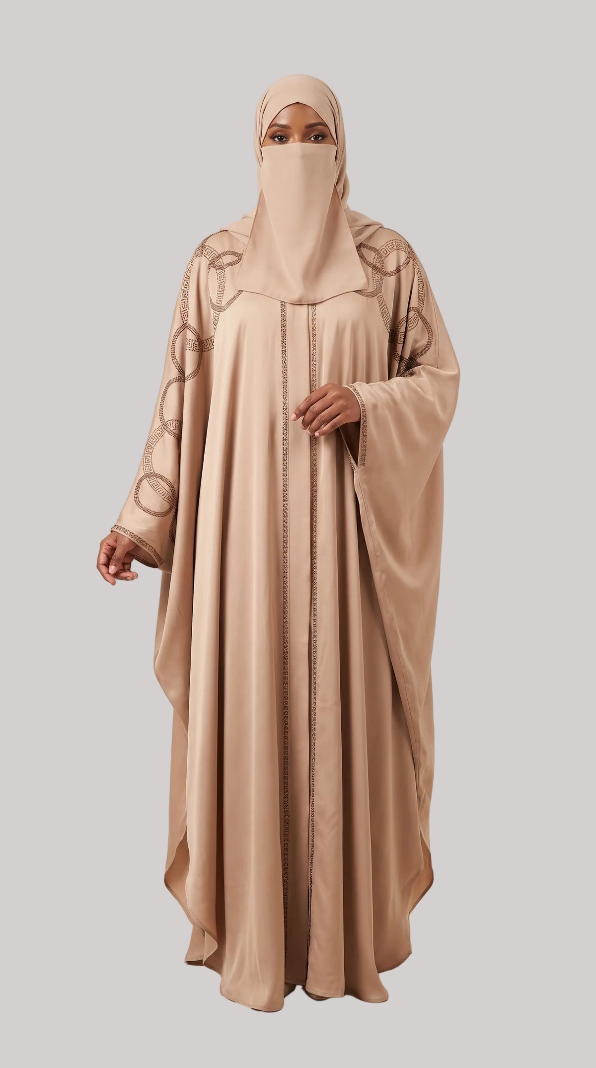 A woman modeling a modest abaya and matching hijab set in a warm beige/tan shade, standing against a plain light beige studio background. The abaya has long flowing sleeves and subtle circular embroidered motifs near the upper sleeves, with a front closure.