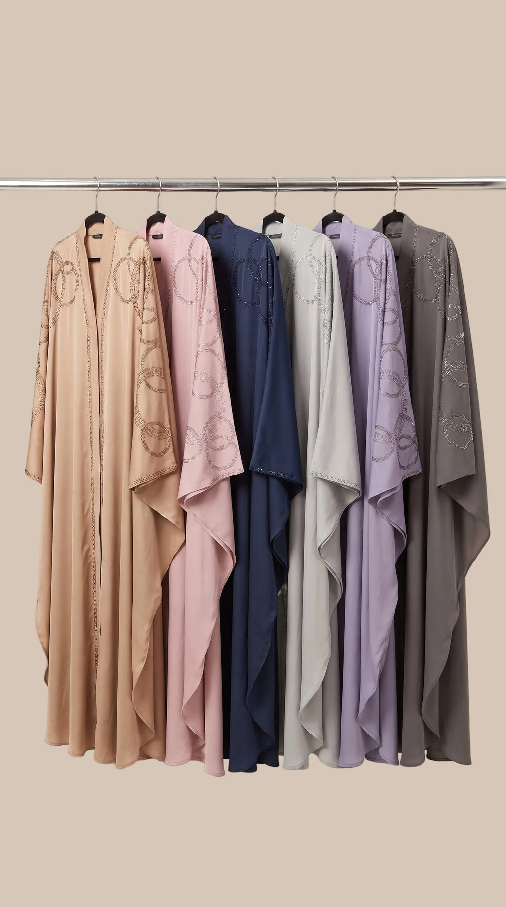 Six lightweight draped kaftan-style abayas in beige, blush pink, navy, light gray, lavender, and charcoal gray hang on a metal rack against a plain off-white background. The beige abaya on the left features a subtle circular pattern.