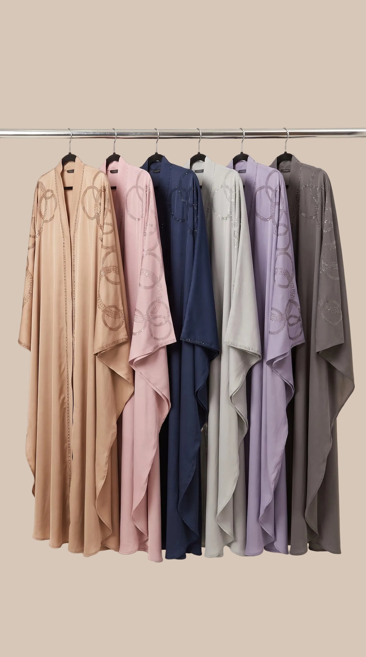 Six lightweight draped kaftan-style abayas in beige, blush pink, navy, light gray, lavender, and charcoal gray hang on a metal rack against a plain off-white background. The beige abaya on the left features a subtle circular pattern.