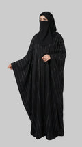 A woman wearing a black abaya with thin vertical metallic stripes and a matching black face covering, standing against a light gray studio background. The brand name LC Abayat  is printed in the top left corner.