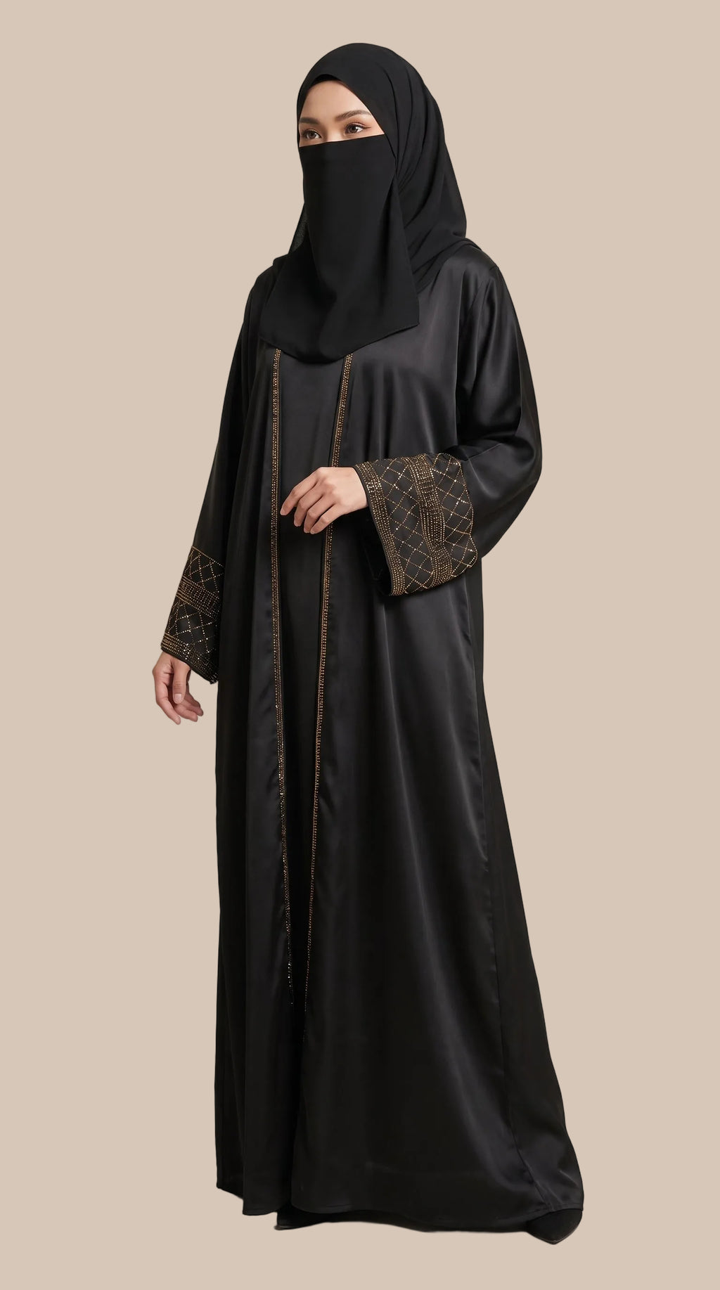 A woman wearing a black abaya with brown patterned sleeve cuffs and a matching black hijab, standing against a plain beige studio background. 
