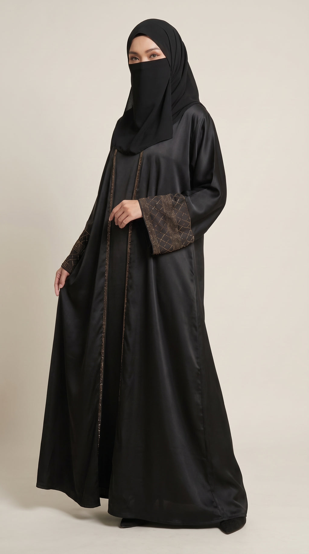 A woman wearing a black abaya with brown patterned sleeve cuffs and a matching black hijab, standing against a plain light beige studio background.