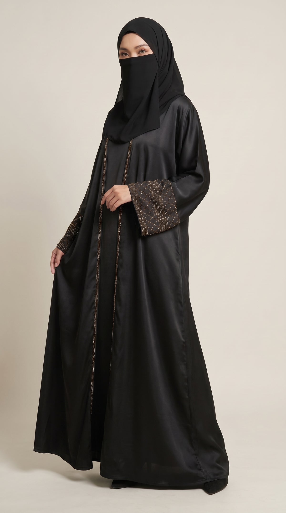 A woman wearing a black abaya with brown patterned sleeve cuffs and a matching black hijab, standing against a plain light beige studio background.