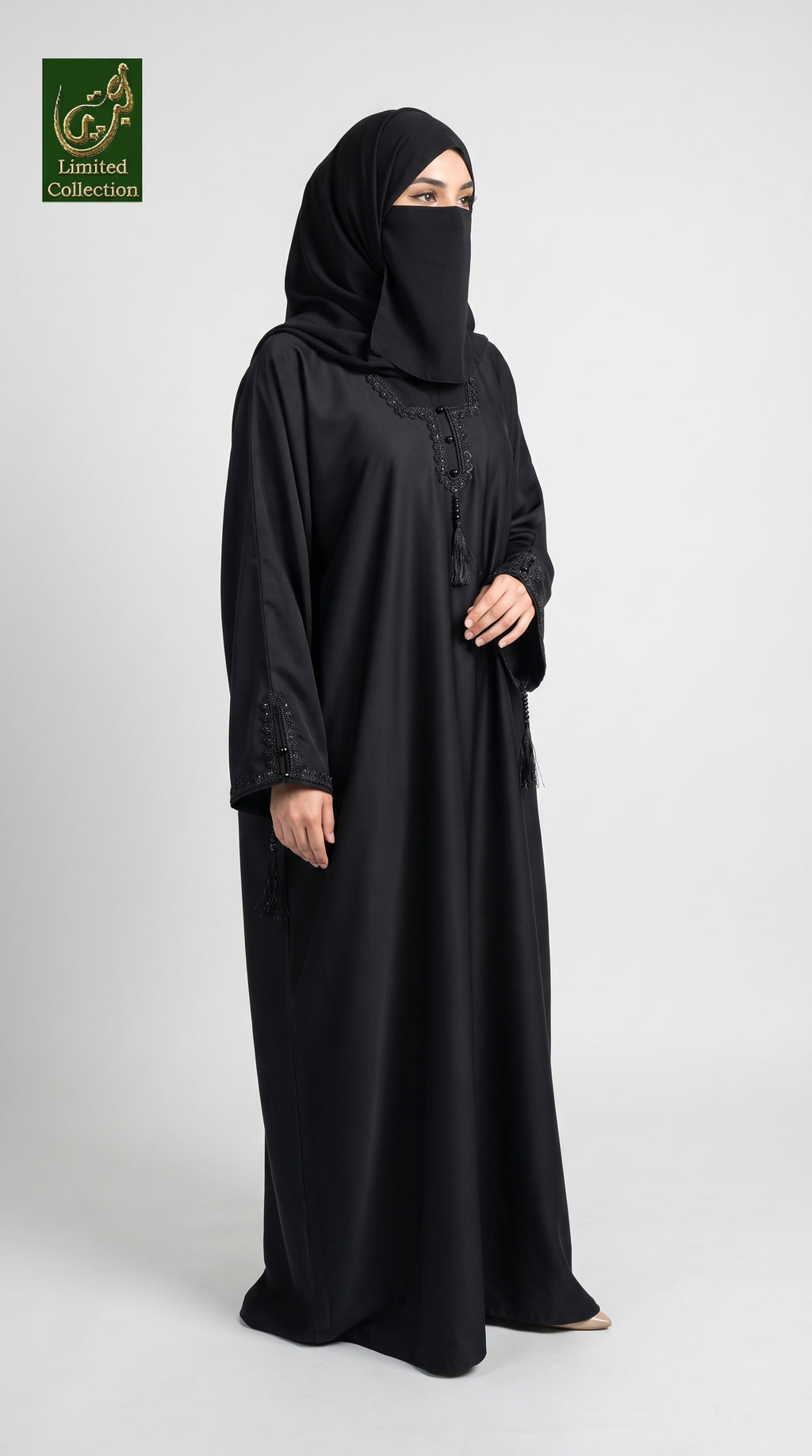 A woman wearing a black abaya and matching hijab stands against a light gray studio background. The abaya has subtle embroidered detailing near the neckline and cuffs, and a small green 'Limited Collection' logo is visible in the top-left corner.
