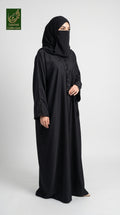 A woman wearing a black abaya and matching hijab stands against a light gray studio background. The abaya has subtle embroidered detailing near the neckline and cuffs, and a small green 'Limited Collection' logo is visible in the top-left corner.