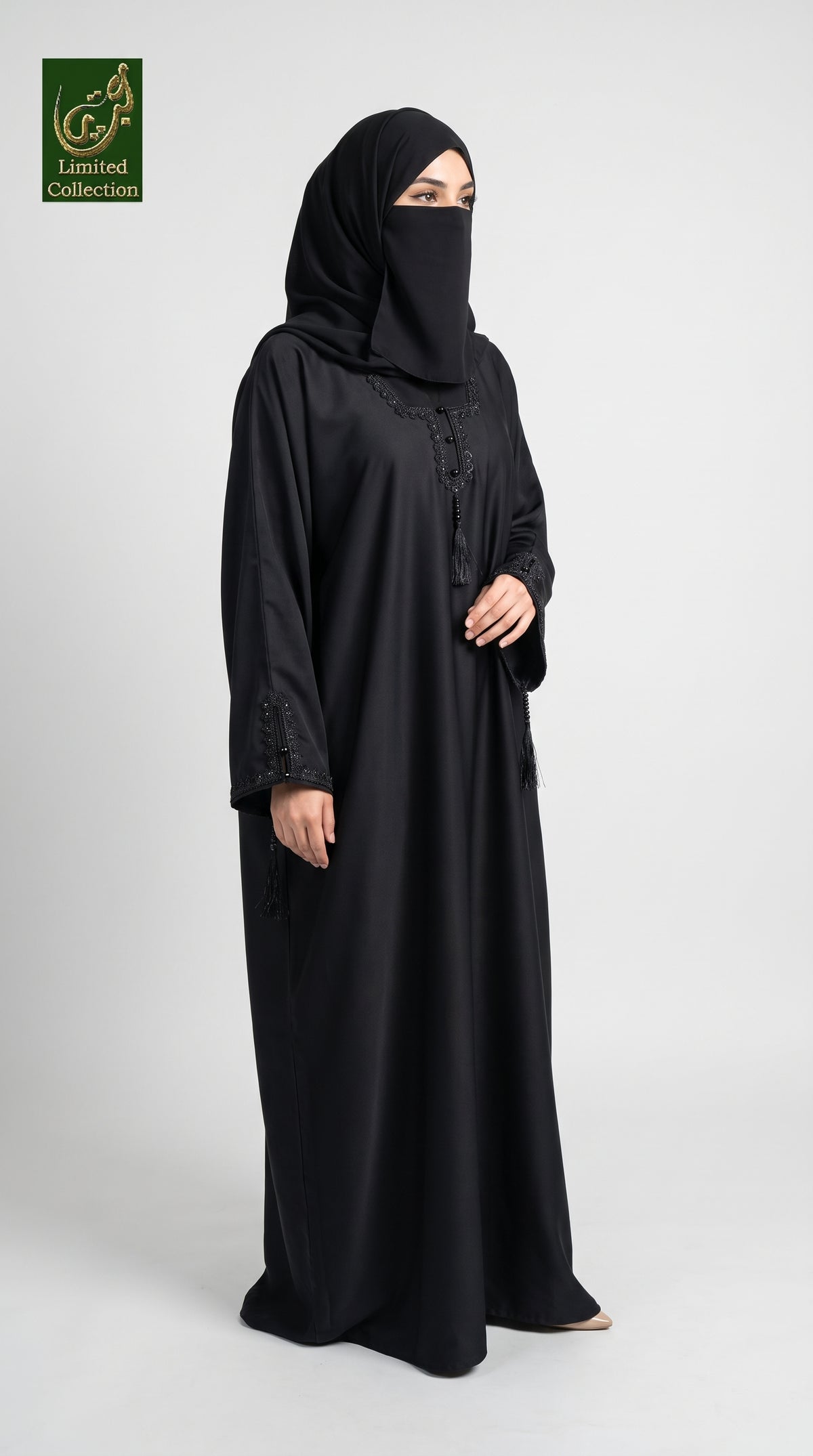 A woman wearing a black abaya and matching hijab stands against a light gray studio background. The abaya has subtle embroidered detailing near the neckline and cuffs, and a small green 'Limited Collection' logo is visible in the top-left corner.