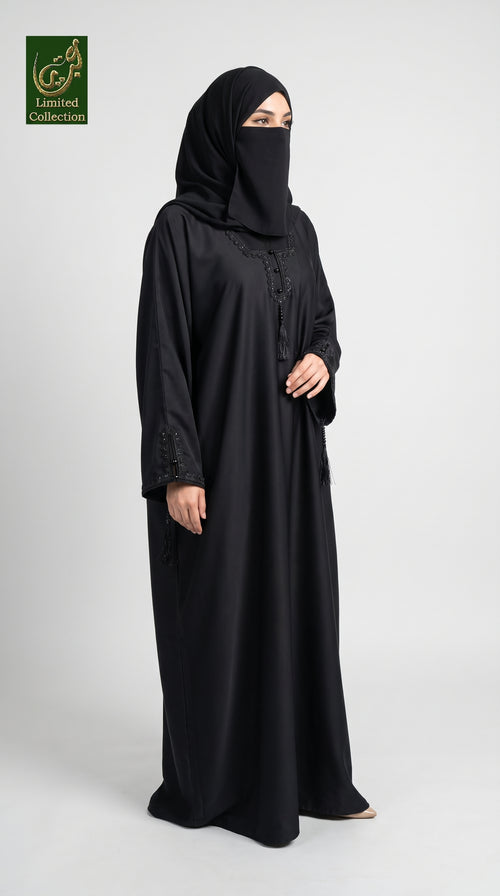 A woman wearing a black abaya and matching hijab stands against a light gray studio background. The abaya has subtle embroidered detailing near the neckline and cuffs, and a small green 'Limited Collection' logo is visible in the top-left corner.