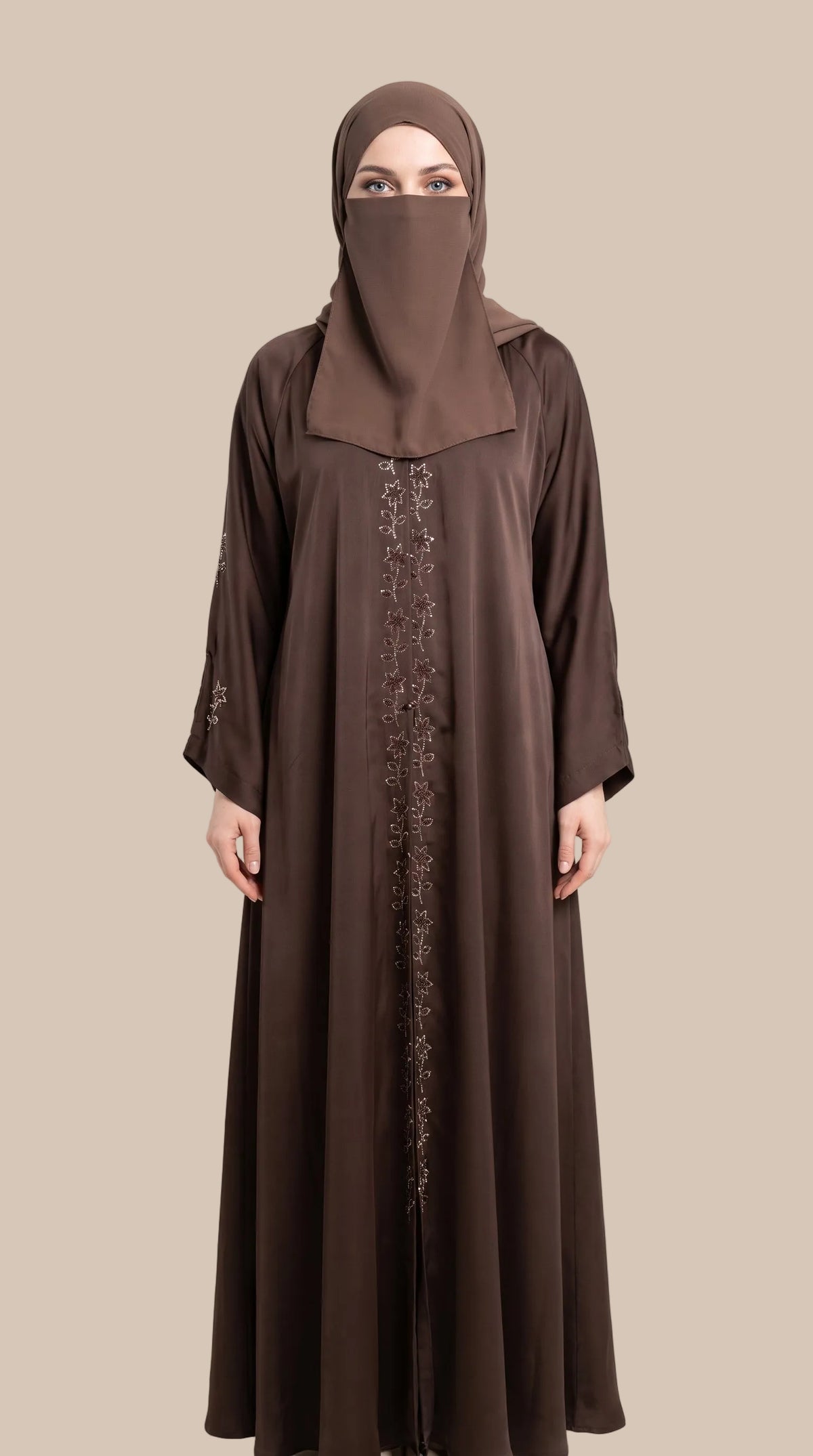 A woman wearing a long brown stonework abaya dress with a matching hijab and a face veil, standing against a plain light beige studio background.