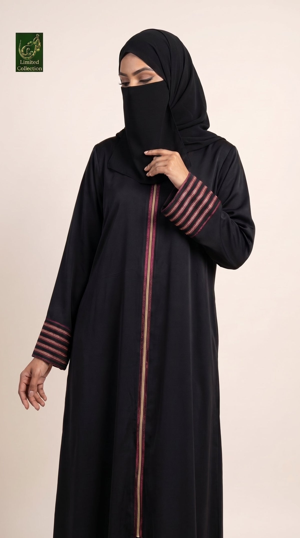 A woman wearing a long black abaya with pink striped cuffs and a vertical pink zipper detail, paired with a matching black hijab, against a solid light beige background.