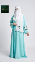 A woman modeling a modest outfit consisting of a long flowing abaya in light aqua blue, a white hijab, and a white waist scarf with black floral embroidery. The background is a clean light gray studio backdrop, with a small brand logo in the top-left corner.