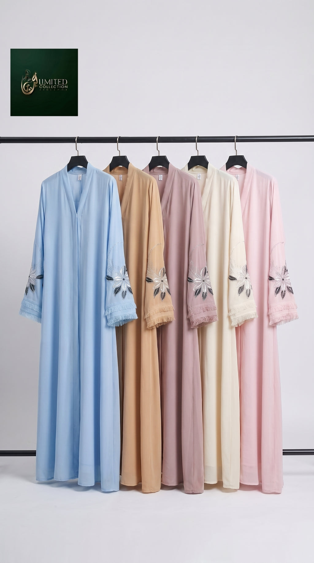 Five long flowing kaftan-style robes hang on a black clothing rack against a plain light gray wall. The robes come in light blue, tan, dusty pink, cream, and soft pink, each with floral embroidery near the sleeve cuffs.
