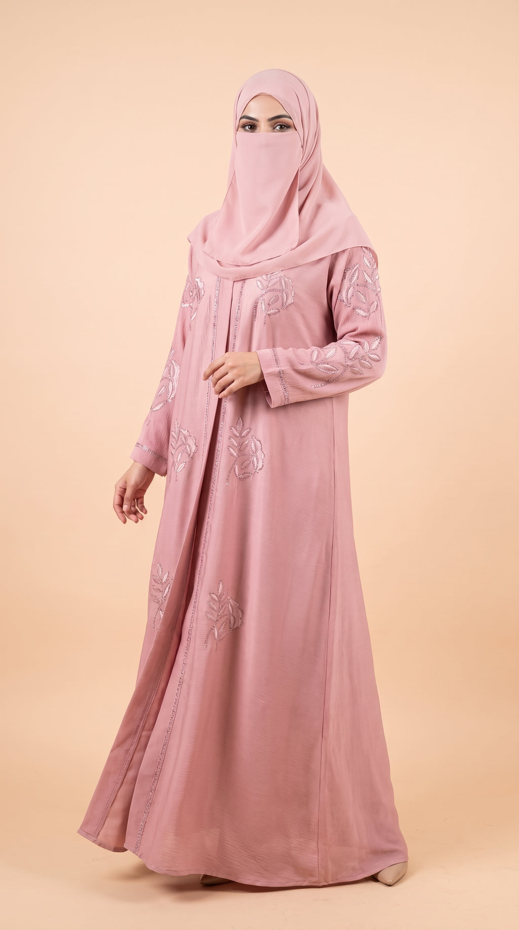 A woman wearing a long dusty pink abaya with floral embroidery and a matching pink hijab, standing against a solid beige studio background.