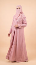 A woman wearing a long dusty pink abaya with floral embroidery and a matching pink hijab, standing against a solid beige studio background.