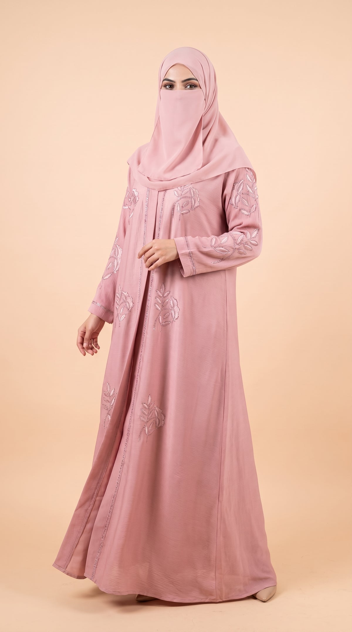 A woman wearing a long dusty pink abaya with floral embroidery and a matching pink hijab, standing against a solid beige studio background.