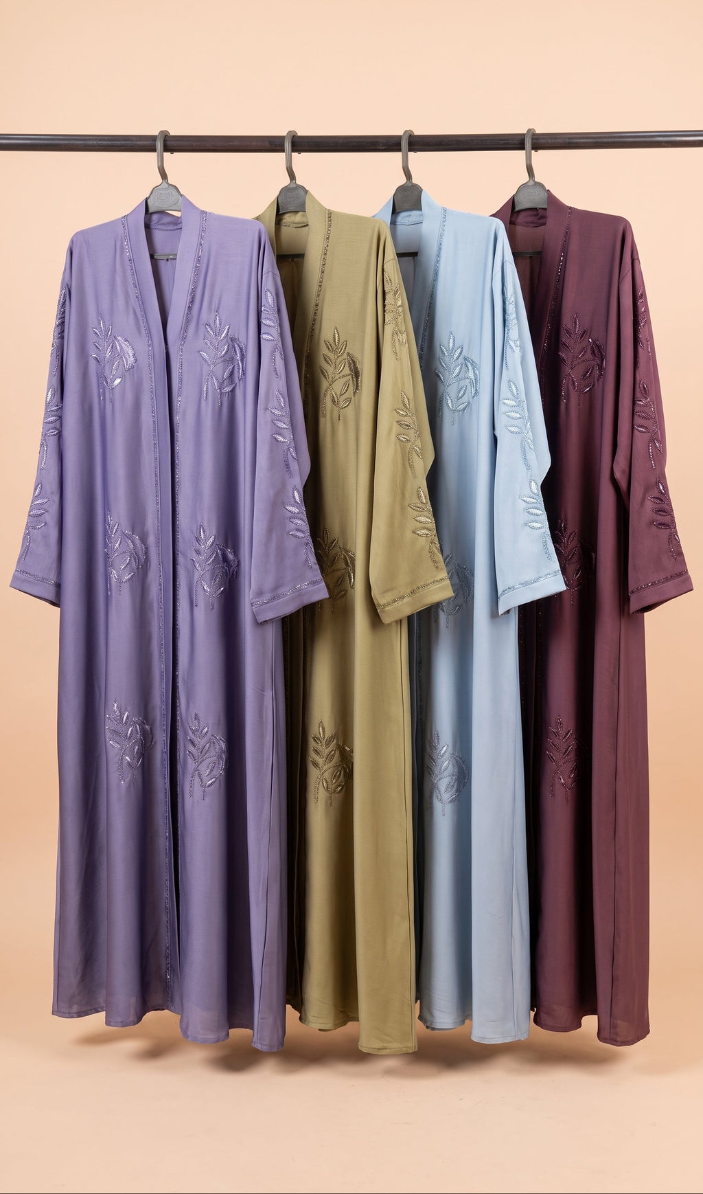 Four long open-front kaftan-style robes hang on a black rod against a warm beige background. From left to right, the robes are lavender purple, olive green, light blue, and plum purple, each with tonal embroidered floral motifs.