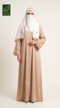 A woman wearing a long beige abaya dress with embroidered sleeves, a white hijab, and a matching headscarf with gold embroidery, standing against a plain light beige studio background. A green 'Limited Collection' logo appears in the top-left corner.