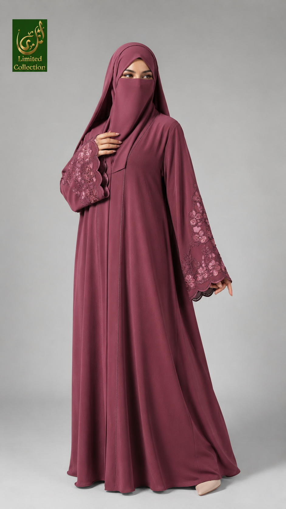 A woman wearing a long, flowing abaya in a dusty rose or mauve color with floral lace detailing on the sleeves, paired with a matching hijab. She is standing against a solid gray studio background.