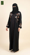 A woman wearing a black abaya with floral embroidery on the chest and sleeves, paired with a matching black niqab, standing against a solid beige background.