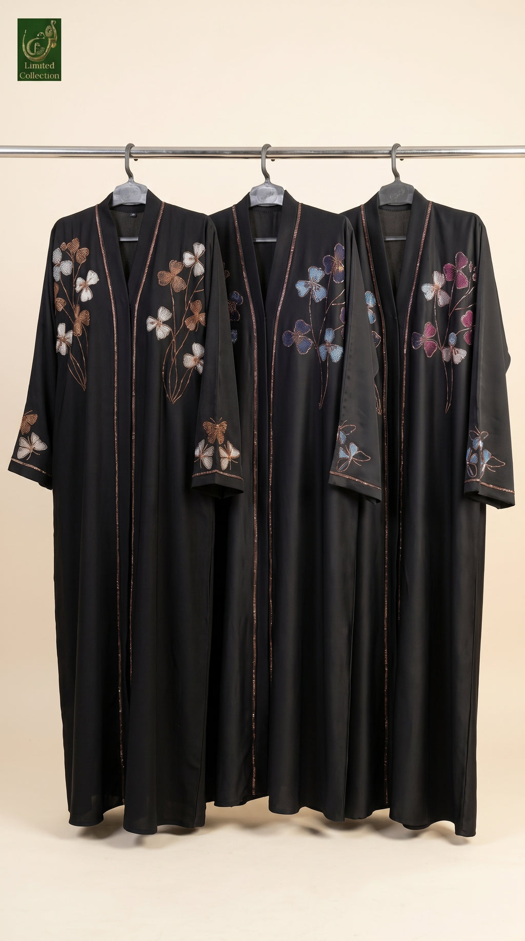 Three black stonework embroidery abayas with colorful floral embroidery hang on a metal rack against a plain beige background. A green brand logo is visible in the top-left corner.