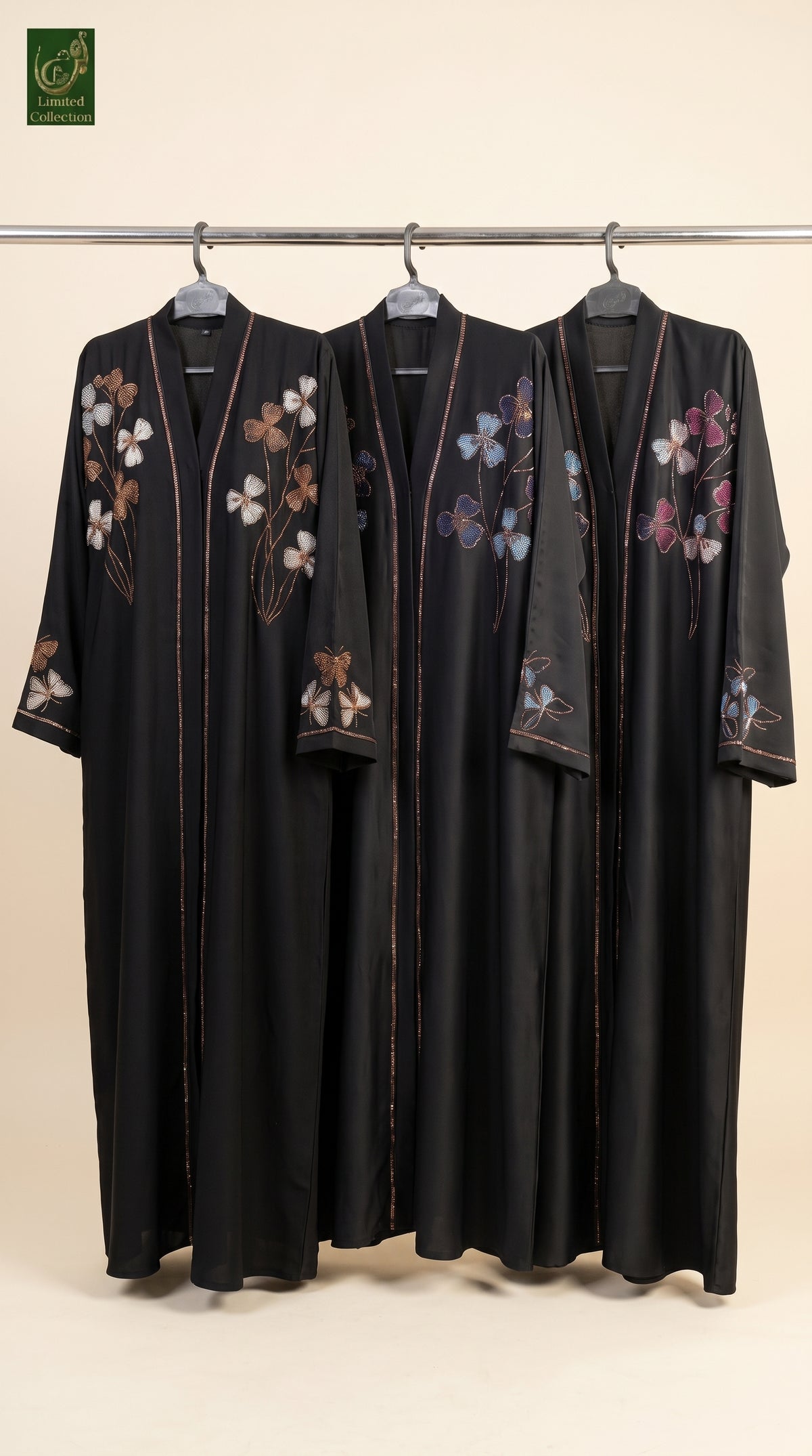 Three black stonework embroidery abayas with colorful floral embroidery hang on a metal rack against a plain beige background. A green brand logo is visible in the top-left corner.