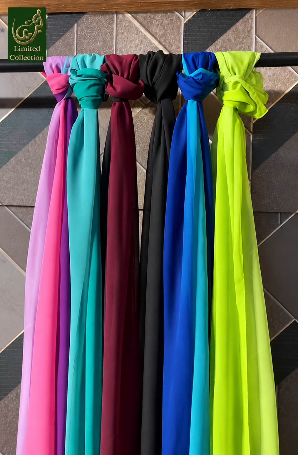 Six lightweight fabric scarves or wraps in different colors are tied and hanging side by side on a black rod. The colors visible are pink, teal, burgundy, black, royal blue, and neon yellow-green.