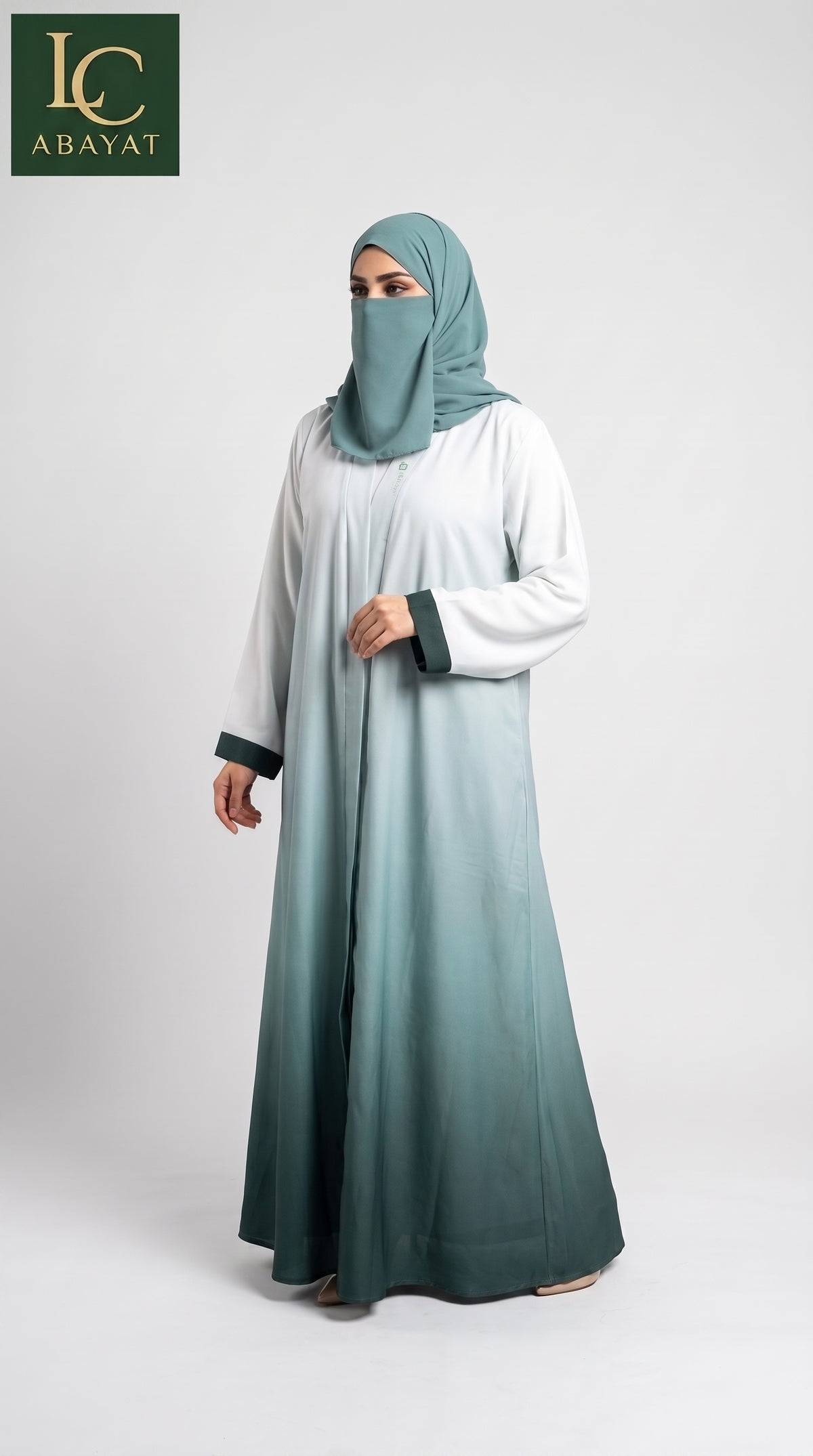 A woman wearing a long front open flowing abaya with a matching hijab, shown against a plain light gray studio background. The abaya has a gradient color story, transitioning from white at the top to a muted teal-green at the bottom, with dark green trim at the cuffs.