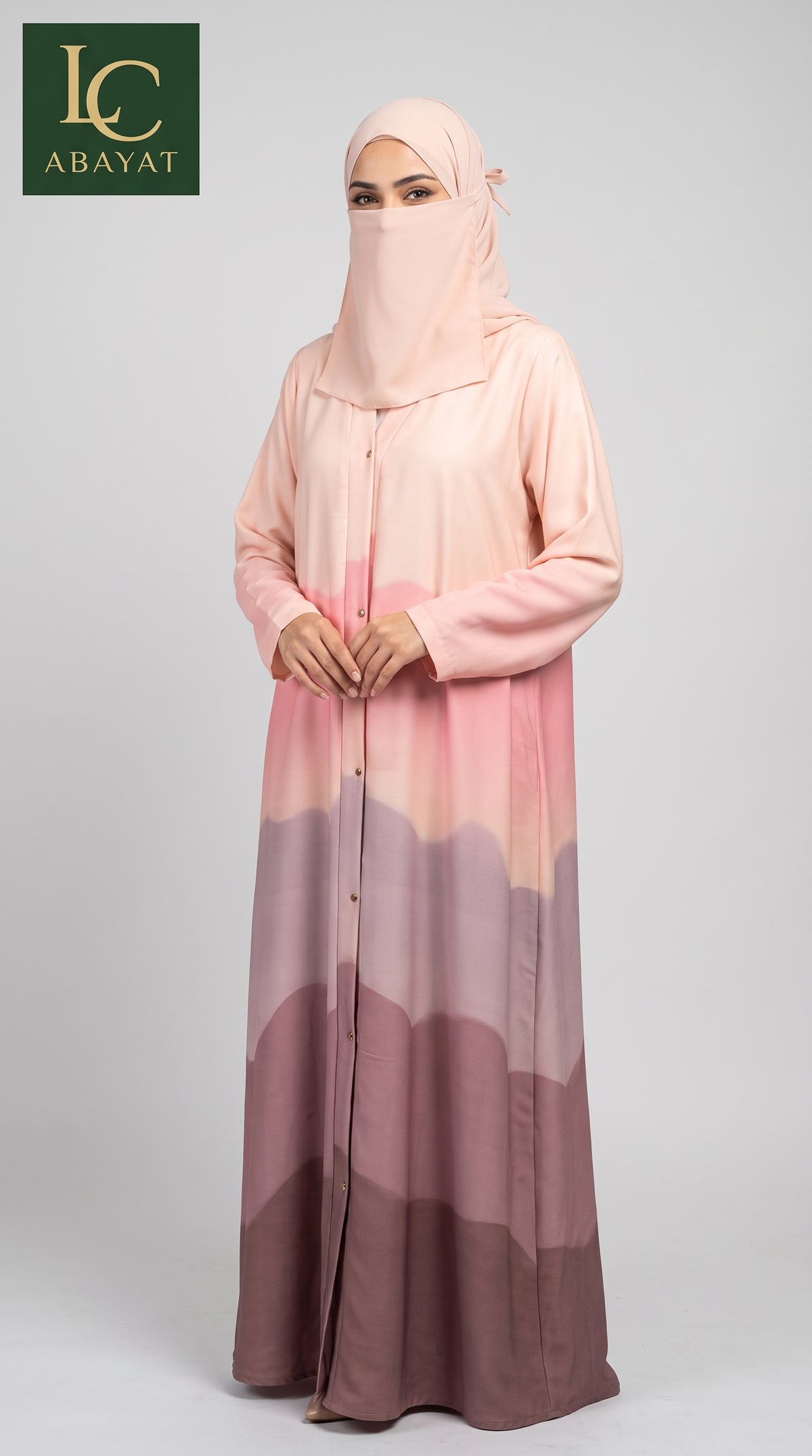 A woman modeling a long abaya dress with a matching hijab, featuring a vertical gradient pattern in shades of pink, peach, and dusty mauve, set against a plain light gray studio background. The 'LC ABAYAT' brand logo is visible in the top-left corner.