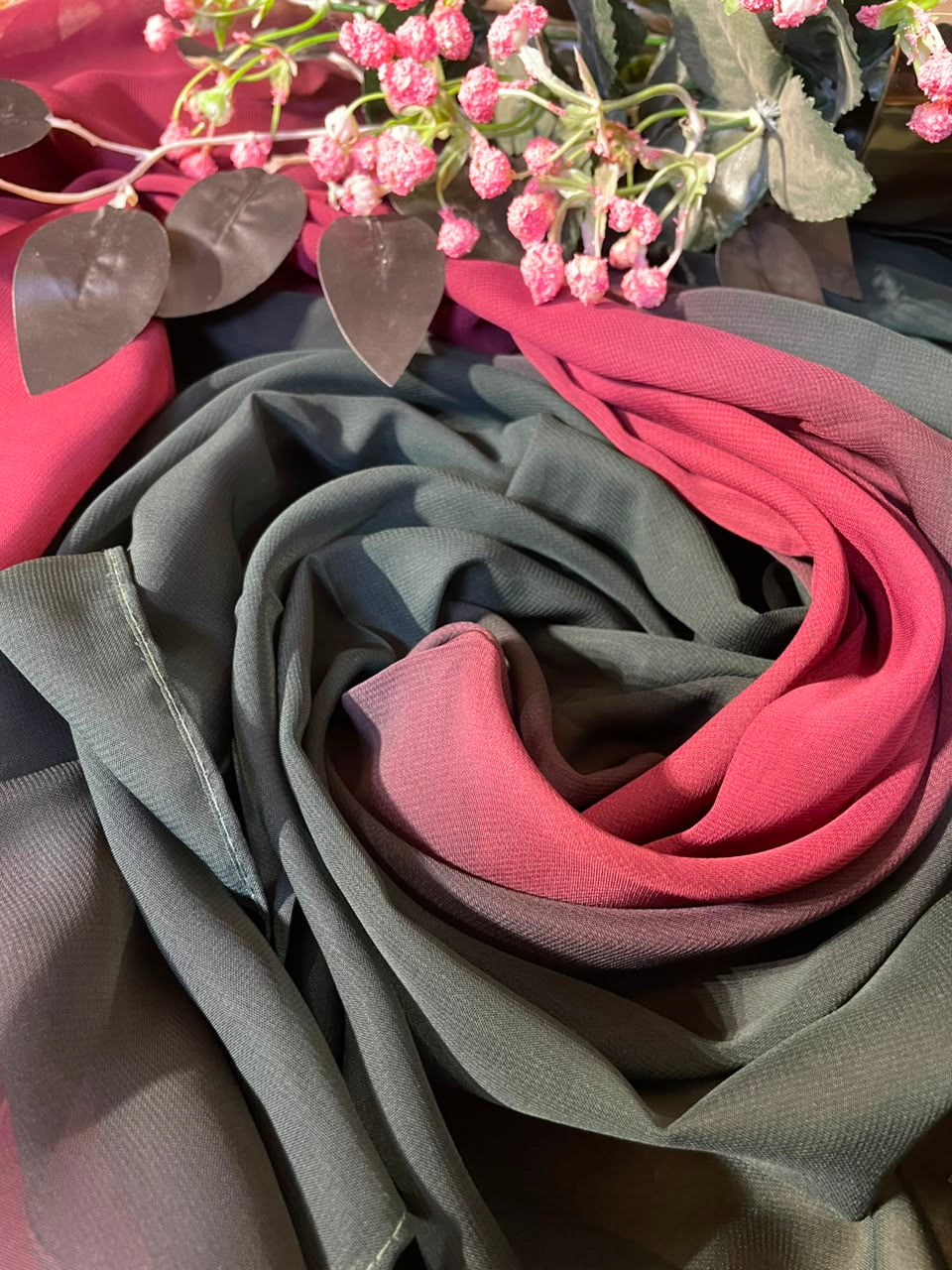 A draped scarf or fabric piece with a color-blocked design, featuring a dark grey outer section, a muted red-pink middle section, and a brighter dusty rose inner section, styled with pink flowers and green leaves on a dark background.