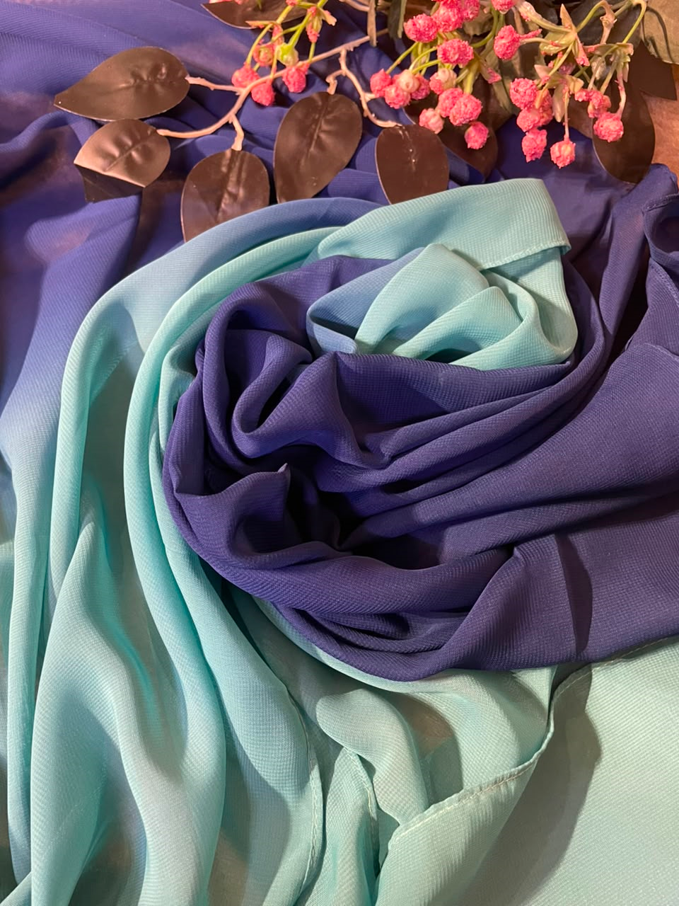 A draped scarf or fabric piece with a smooth gradient transitioning from light mint green to medium blue and finally to a rich purple, styled with pink flowers and metallic leaves on a matching purple background.