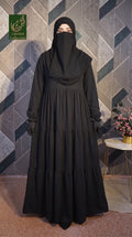 A woman wearing a long black modest Tiered Frill Maxi Abaya with a full face veil and black gloves, standing in front of a geometric gray tile wall. A small side table with flowers is visible on the right, and a green 'Limited Collection' logo appears in the top-left corner.