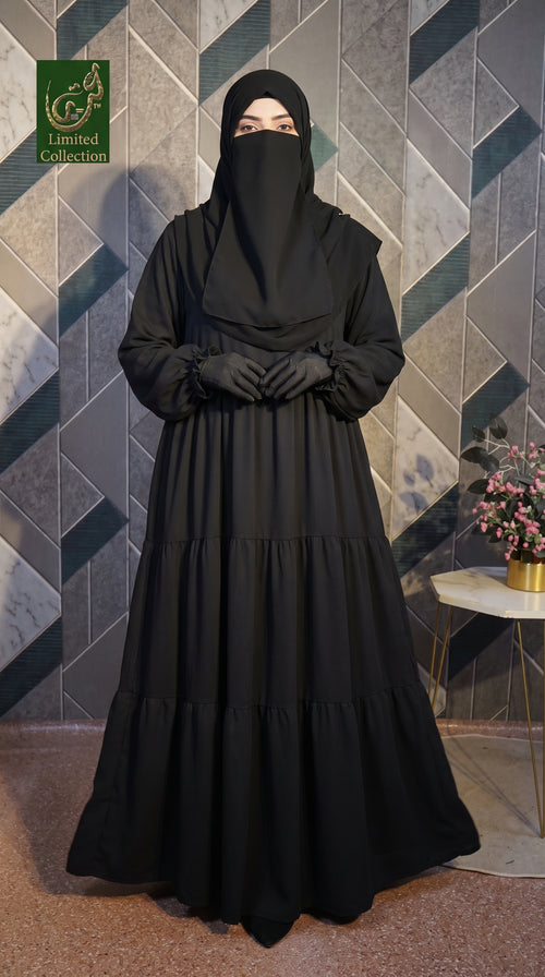 A woman wearing a long black modest Tiered Frill Maxi Abaya with a matching face veil, black gloves, and a black head covering, standing in front of a geometric gray tile wall. A small side table with flowers is visible to the right, and a green 'Limited Collection' logo appears in the top-left corner.