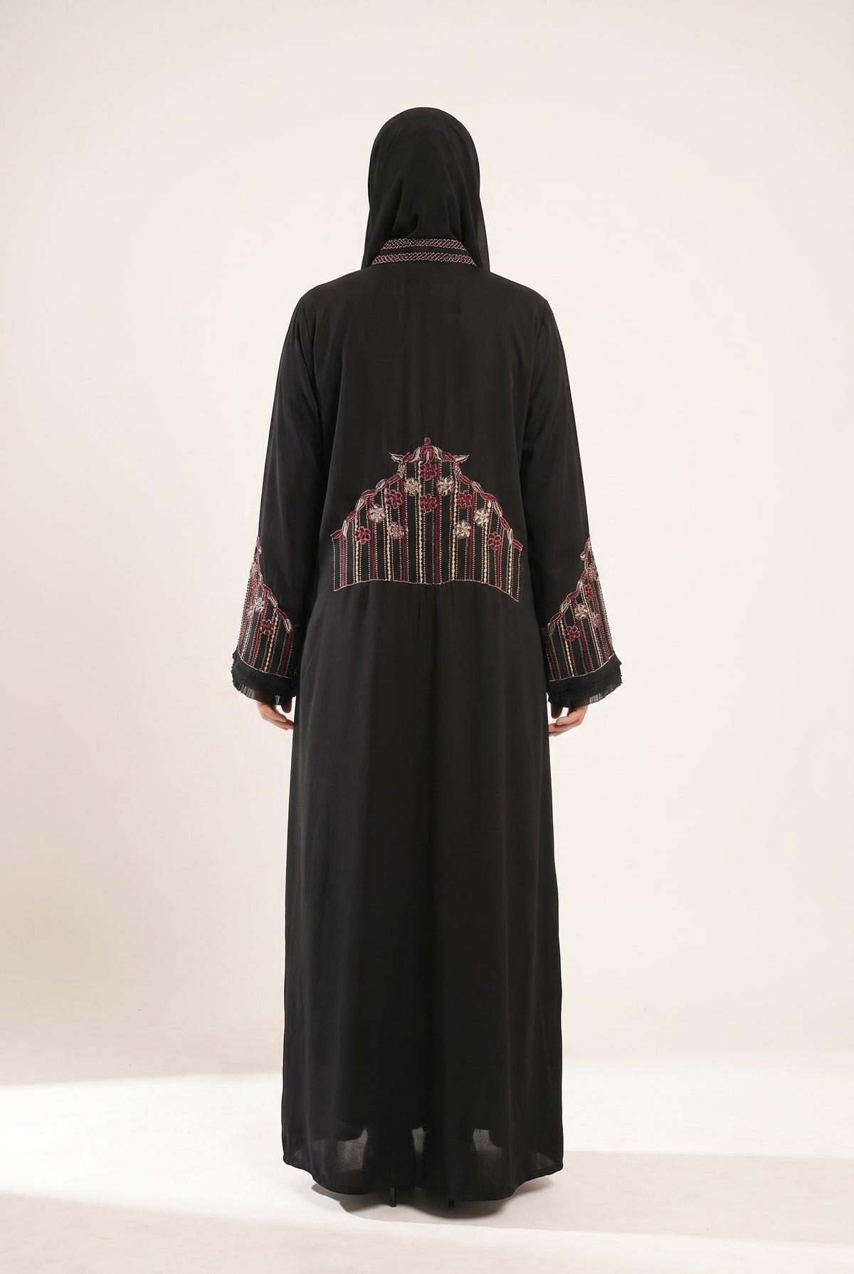 A woman viewed from behind wearing a long black abaya with intricate multicolor motif crown embroidery on the back and sleeve cuffs, paired with a matching black headscarf, set against a plain off-white studio background.