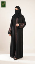 A woman modeling a long black abaya with red and green patterned sleeve cuffs and a vertical red stripe detail down the front, paired with a matching black headscarf and face veil, against a plain off-white studio background.