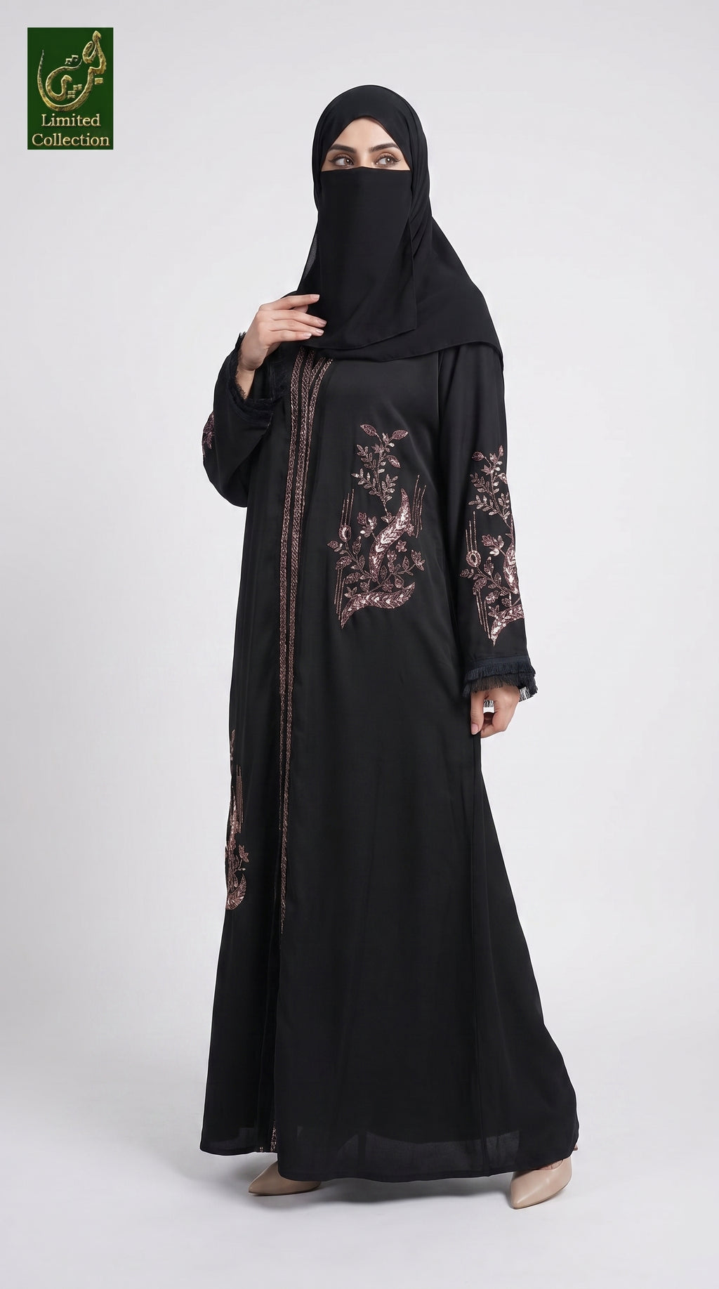 A woman wearing a black abaya with rose gold floral embroidery on the chest and sleeves, paired with a matching black hijab. She is standing against a light gray studio background.