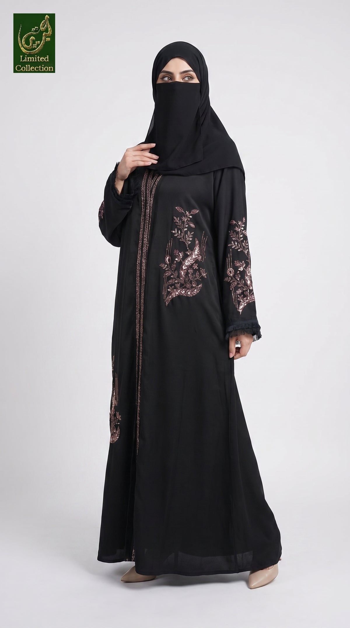 A woman wearing a black abaya with rose gold floral embroidery on the chest and sleeves, paired with a matching black hijab. She is standing against a light gray studio background.