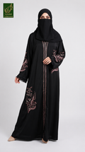 A woman wearing a black abaya with pink floral embroidery and a matching black hijab, standing against a light gray studio background. A green 'Abayat Limitef Collection' logo is visible in the top-left corner.