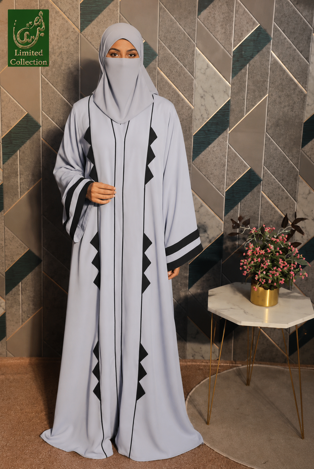 A woman wearing a light gray cutwork abaya with black geometric triangle patterns and a matching gray hijab, standing in front of a geometric tiled wall next to a small table with flowers. A green 'Limited Collection' logo is visible in the top left corner.