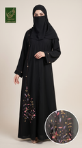 A woman modeling a long black abaya with a matching black hijab, shown against a plain beige studio background. The abaya has delicate gold bead trim and colorful floral embroidery near the lower front, with a circular inset close-up showing the embroidery detail.