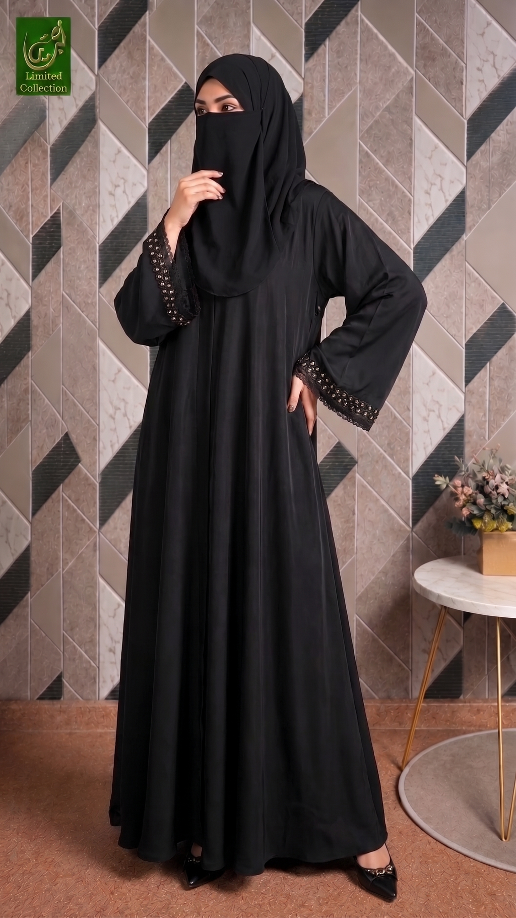 A woman wearing a black abaya and matching hijab with decorative gold embellishments on the sleeve cuffs, standing in front of a geometric patterned wall.