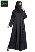 A woman modeling a black full-length abaya with matching hijab and black shoes against a plain white background. The abaya features decorative black embroidered patterns near the lower hem and sleeve cuffs.