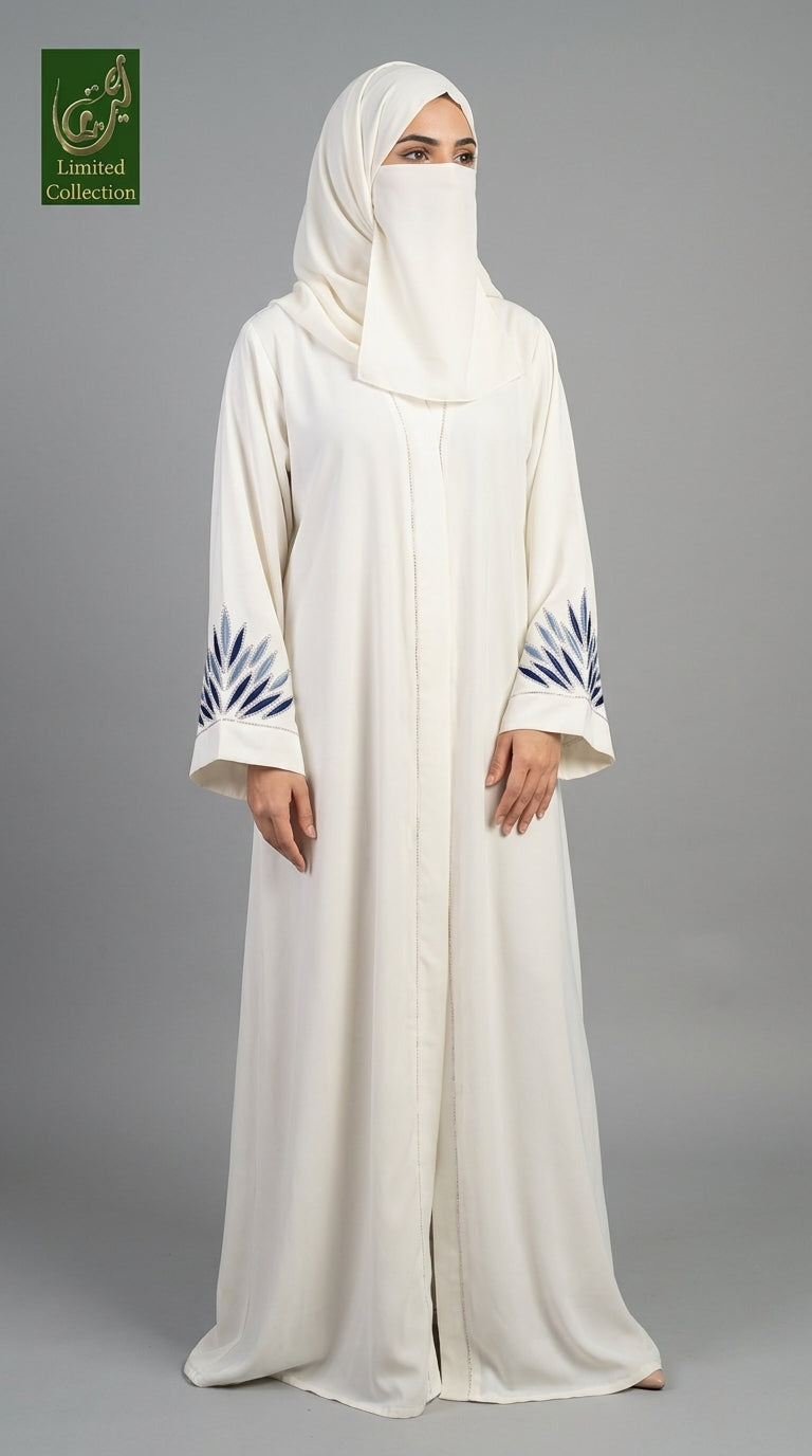 A woman wearing a long white abaya with blue leaf embroidery on the cuffs and a matching white hijab, standing against a solid gray studio background. A green 'Limited Collection' logo is visible in the top-left corner.