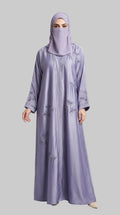 A woman wearing a long lavender abaya with subtle tonal floral embroidery and a matching hijab, standing against a light gray studio background.