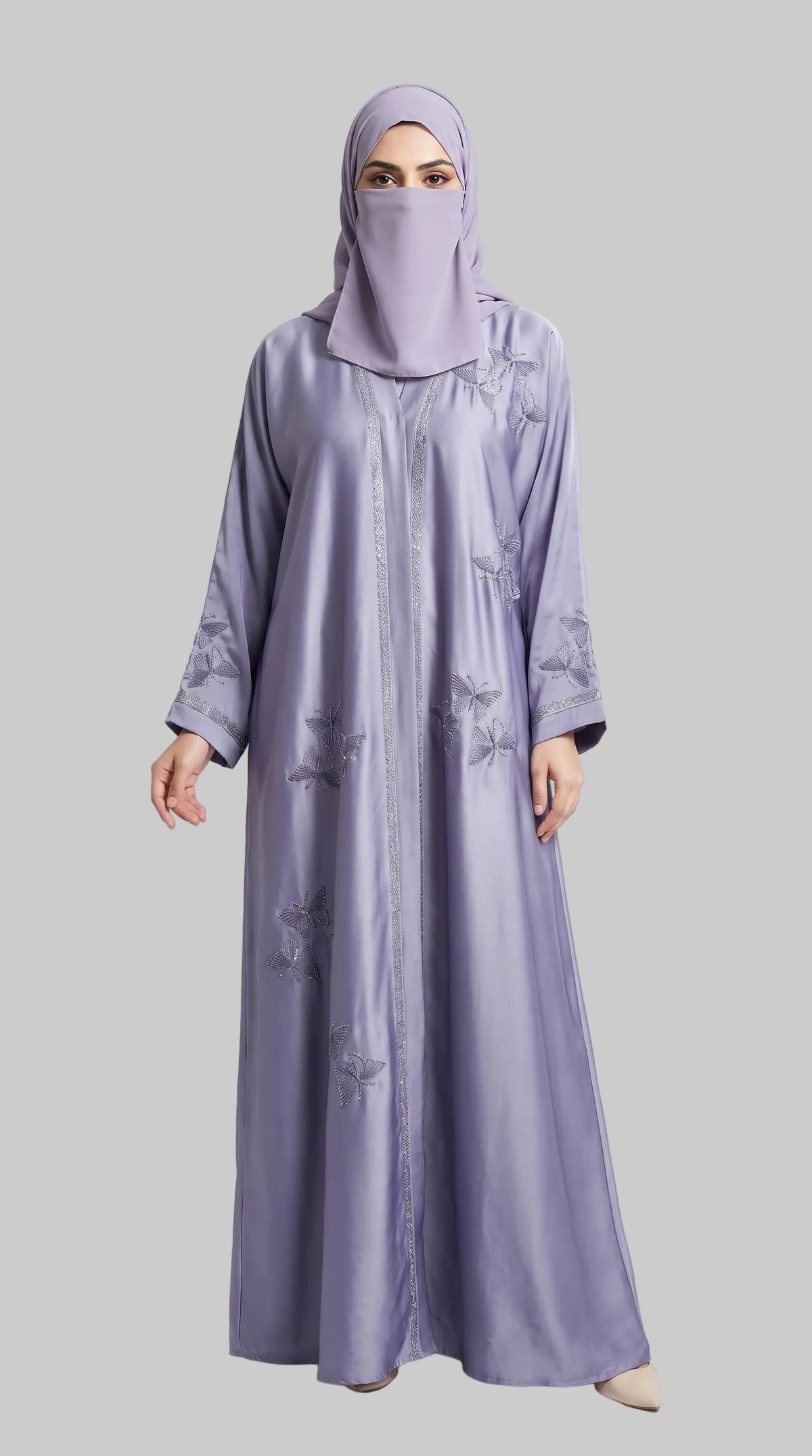 A woman wearing a long lavender abaya with subtle tonal floral embroidery and a matching hijab, standing against a light gray studio background.