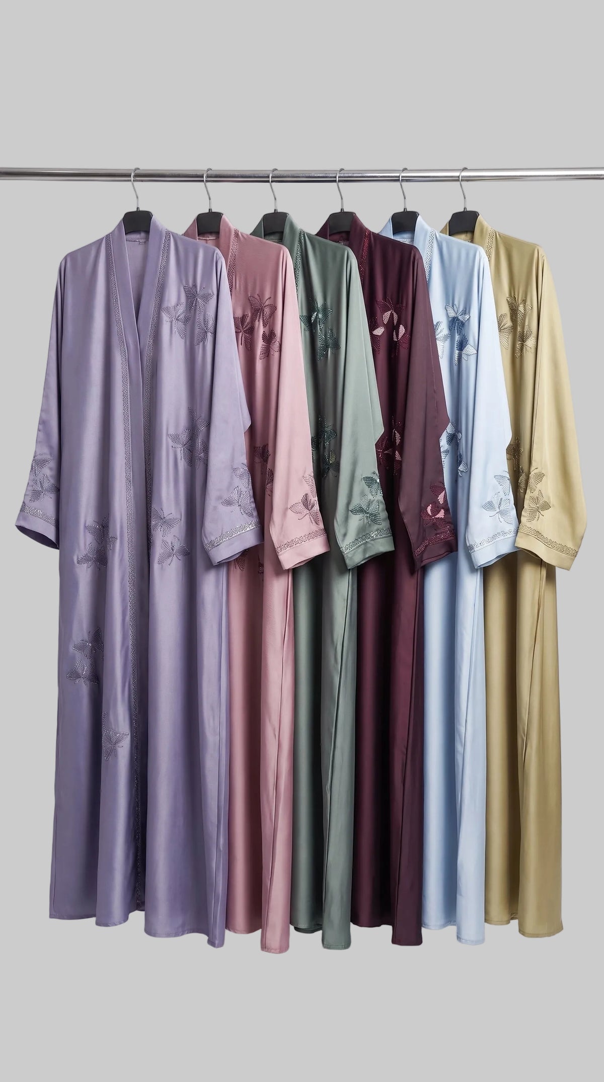 Six long stonework embroidery abayas in different colors hang on a metal rack against a light gray studio background. The colors visible are lavender, dusty pink, sage green, burgundy, light blue, and mustard gold.