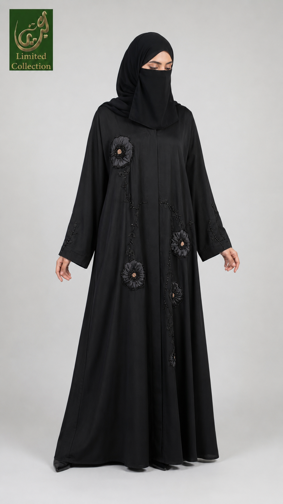 A woman wearing a long black abaya with matching black hijab, standing against a light gray studio background. The abaya features black floral applique details on the front.